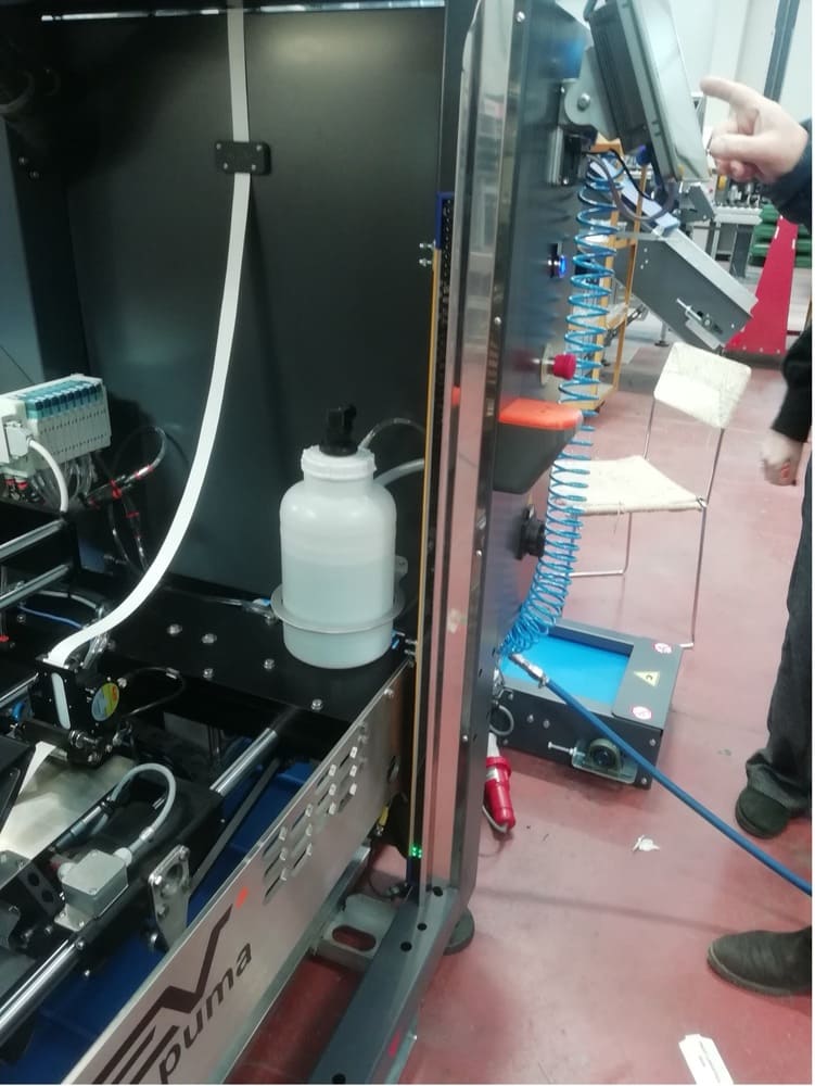 Vista laterale di un macchinario industriale durante un test funzionale. In evidenza il pannello di controllo con touch screen, un tubo a spirale blu, componenti pneumatici interni e la sottile barriera di sicurezza installata verticalmente.